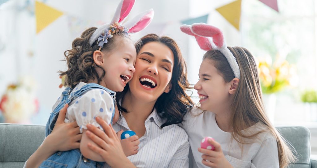 Deux fillettes souriantes et portant des oreilles de lapin entourent leur maman, dans un décor de Pâques.