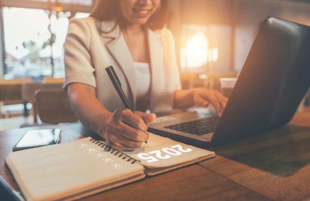 Jeune femme souriante inscrivant dans un carnet ses résolutions du Nouvel An 2025, assise au bureau devant son ordinateur.