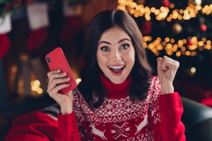 Jeune femme très heureuse, levant les mains en signe de victoire, tenant son cellulaire dans une main dans un décor de Noël.