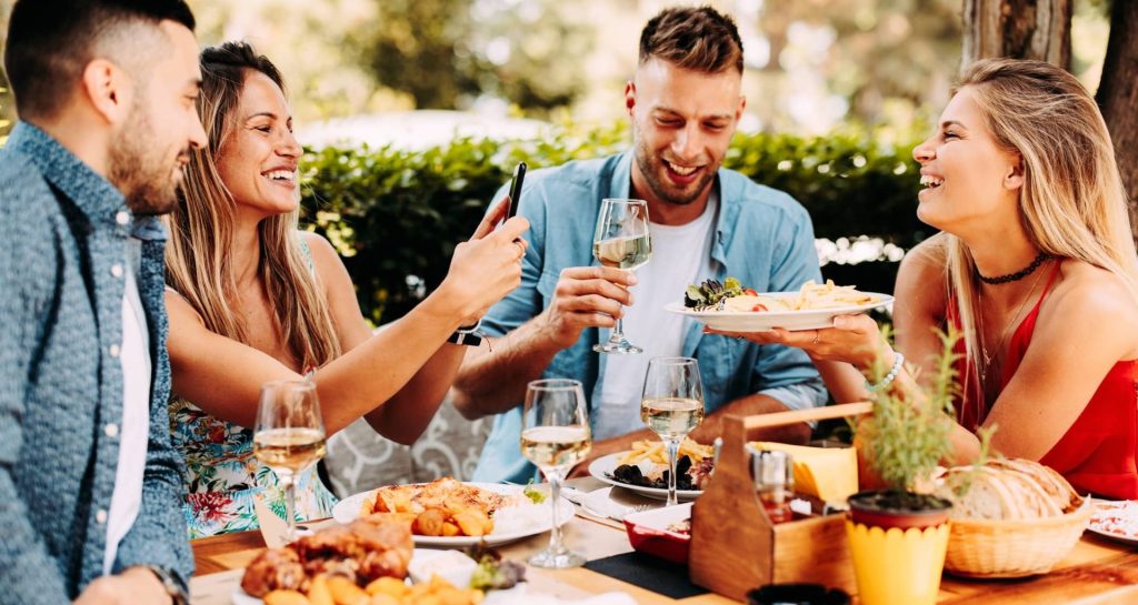 Deux couples d'amis jeunes et souriants attablés en restaurant terrasse, autour d'un copieux repas et de verres de vin.