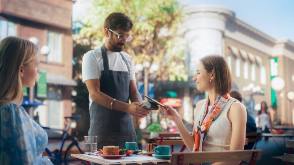 Femme souriante payant au serveur son repas en restaurant terrasse, pris en compagnie d'une amie. 